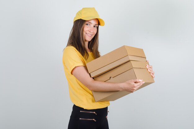 Lieferfrau im gelben T-Shirt, in der Hose, in der Kappe, die Pappkartons hält und froh aussieht