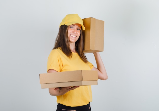 Lieferfrau im gelben T-Shirt, in der Hose, in der Kappe, die Pappkartons hält und lächelt