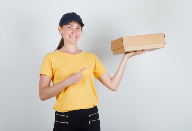 Lieferfrau im T-Shirt, in der Hose, in der Kappe, die Finger auf Pappkarton zeigt und fröhlich schaut