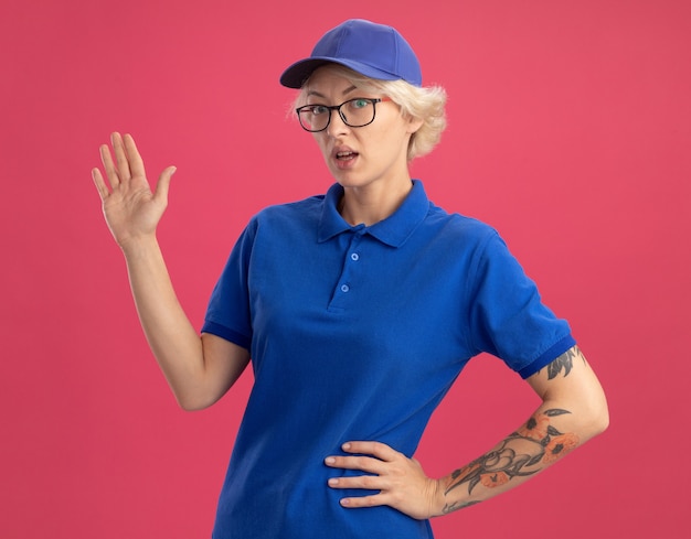 Lieferfrau in blauer uniform und mütze, die verwirrt mit arm oh hand über rosa wand präsentiert