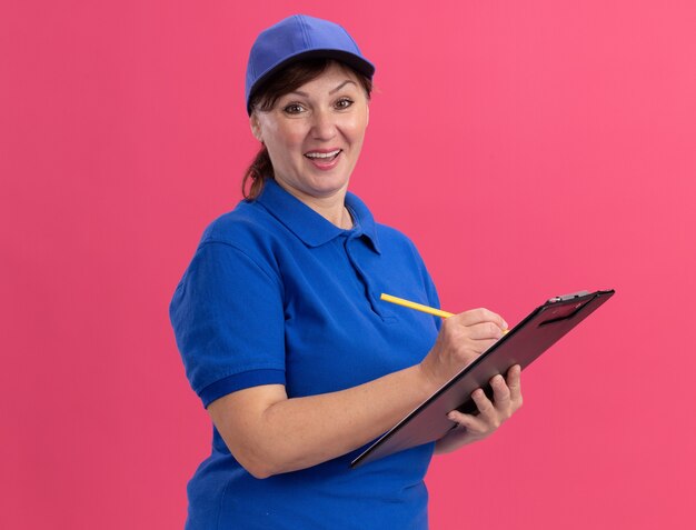 Lieferfrau mittleren Alters in blauer Uniform und Kappe, die Klemmbrett und Bleistiftschrift hält, die Front hapy und positives Lächeln betrachtet, das fröhlich über rosa Wand steht