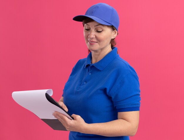 Lieferfrau mittleren Alters in blauer Uniform und Kappe, die Zwischenablage mit leeren Seiten hält, die es mit Lächeln auf Gesicht betrachten, das über rosa Wand steht
