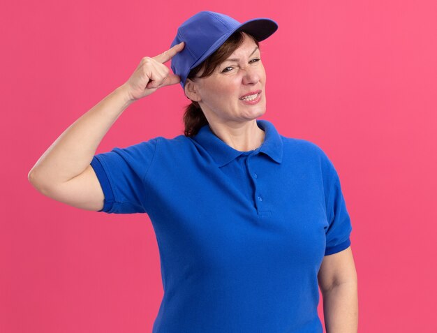 Lieferfrau mittleren Alters in blauer Uniform und Mütze, die verwirrt und sehr besorgt mit der Hand auf ihrem Kopf sieht, der über rosa Wand steht