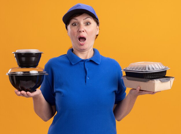 Lieferfrau mittleren Alters in blauer Uniform und Mütze mit Lebensmittelverpackungen, die vorne erstaunt und überrascht über der orangefarbenen Wand stehen