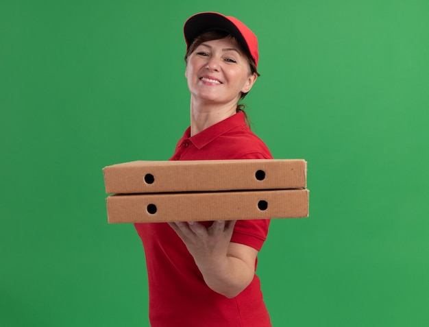 Lieferfrau mittleren Alters in der roten Uniform und in der Kappe, die Pizzaschachteln hält, die vorne lächelnd mit glücklichem Gesicht stehen über grüner Wand stehen