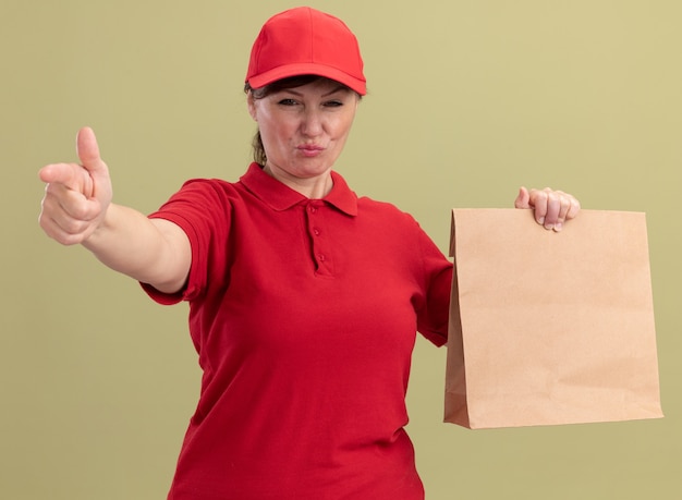 Lieferfrau mittleren Alters in roter Uniform und Kappe, die Papierpaket gibt, das vorne mit ernstem sicherem Ausdruck schaut, der mit Zeigefinger an der Vorderseite steht über grüner Wand