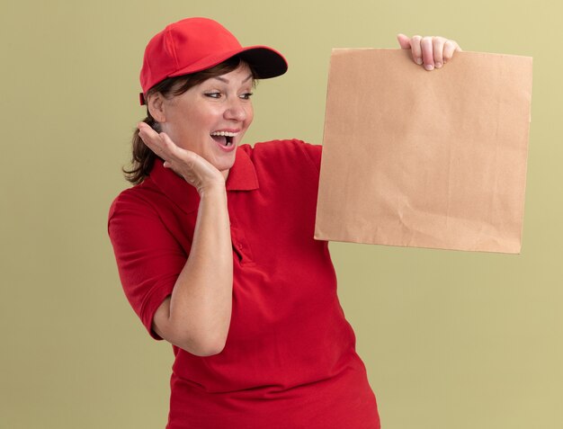 Lieferfrau mittleren Alters in roter Uniform und Kappe, die Papierpaket hält, das es glücklich und aufgeregt betrachtet, über grüner Wand stehend