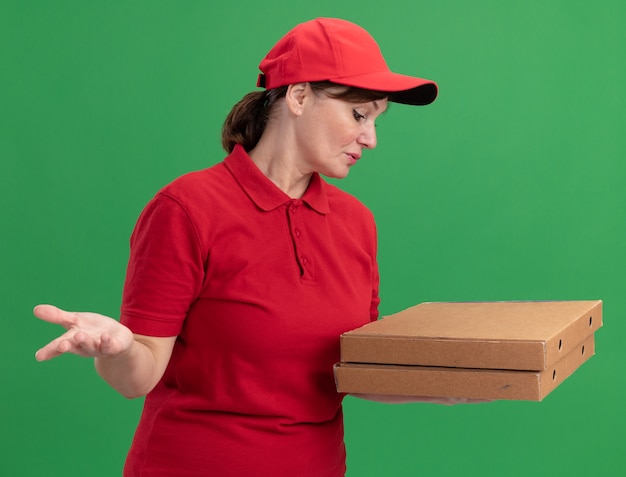 Lieferfrau mittleren Alters in roter Uniform und Kappe, die Pizzaschachteln hält, die sie verwirrt betrachten, die über grüner Wand stehen