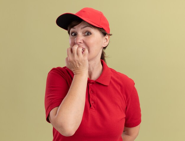 Lieferfrau mittleren Alters in roter Uniform und Mütze, die die gestressten und nervösen Beißnägel der Front betrachtet, die über grüner Wand stehen