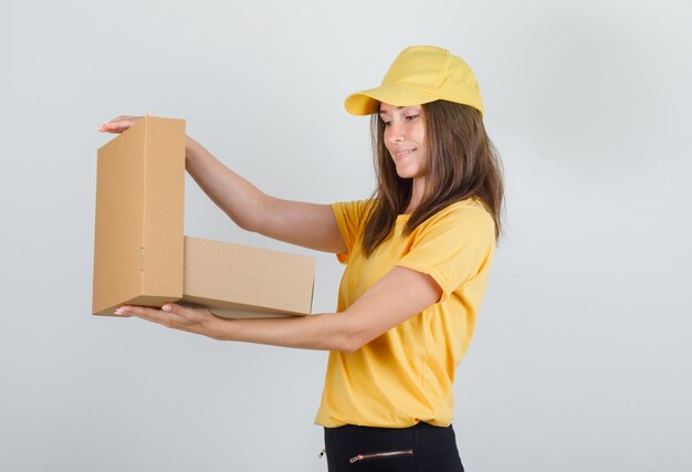 Lieferfrau öffnet Pappkarton in gelbem T-Shirt, Hose, Mütze und sieht froh aus