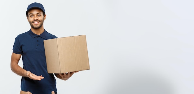 Lieferkonzept Porträt eines glücklichen afroamerikanischen Liefermanns, der mit der Hand zeigt, um ein Kartonpaket zu präsentieren, das auf grauem Studio-Hintergrund-Kopienraum isoliert ist