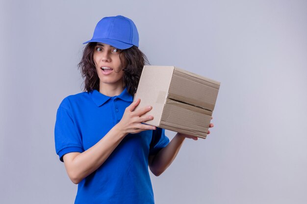 Liefermädchen in der blauen Uniform und in der Kappe, die Boxpaket hält, das überrascht steht, auf lokalisiertem Weiß stehend