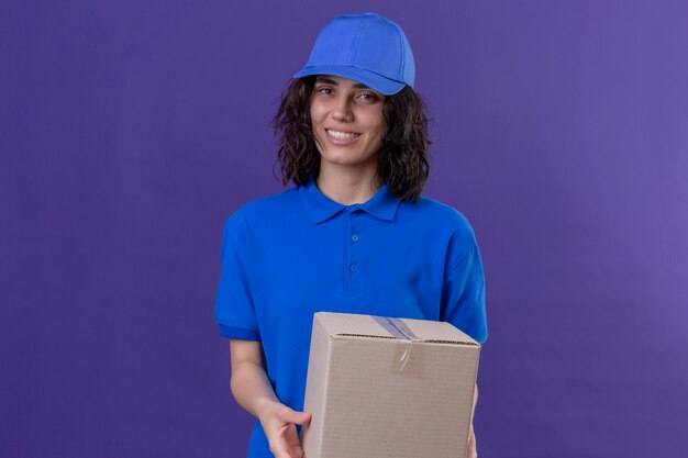 Liefermädchen in der blauen Uniform und in der Kappe, die Boxpaket lächelnd freundlich, positiv und glücklich steht auf lokalisiertem Purpur hält