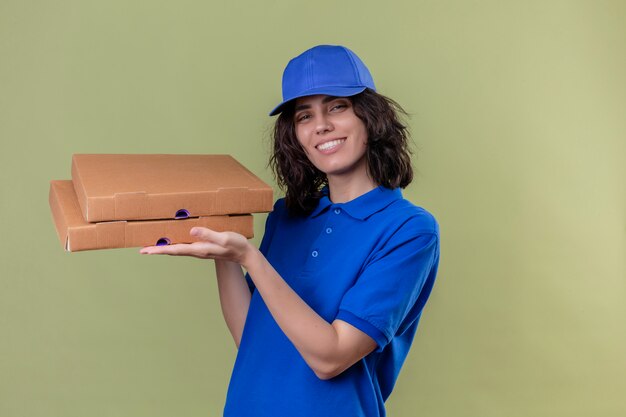 Liefermädchen in der blauen Uniform und in der Kappe, die die freundlichen und selbstbewussten stehenden Pizzaschachteln hält, die auf isolierter olivgrüner Farbe stehen