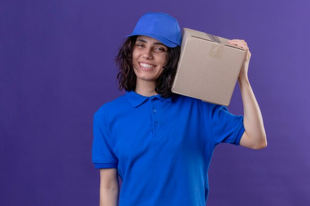 Liefermädchen in der blauen Uniform und in der Kappe, die Kastenpaket auf der Schulter hält, lächelt freundlich positiv und glücklich stehend