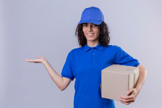 Liefermädchen in der blauen Uniform und in der Kappe, die Kastenpaket darstellt, das mit dem Arm der Hand präsentiert, der fröhlich stehend lächelt