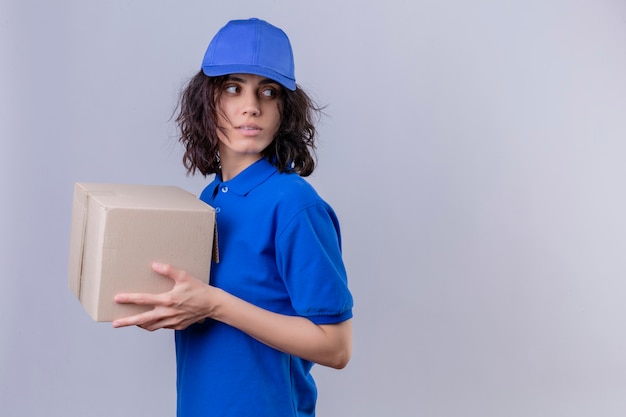 Liefermädchen in der blauen Uniform und in der Kappe, die Kastenpaket hält, das seitlich steht und mit ernstem Gesicht auf Weiß wegschaut