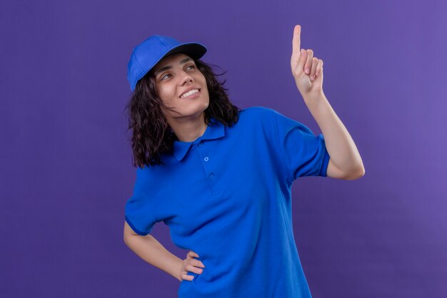 Liefermädchen in der blauen Uniform und in der Kappe, die mit dem Finger oben lächelnd mit dem glücklichen Gesicht stehend zeigt