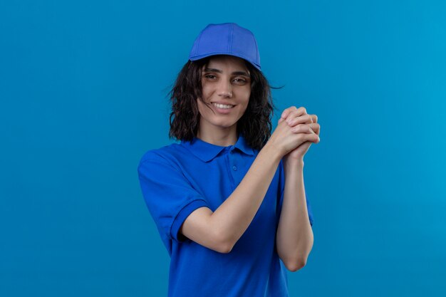 Liefermädchen in der blauen Uniform und in der Kappe, die mit einer Geste der Teamarbeit stehen, die freundlich auf lokalisiertem Blau lächelt