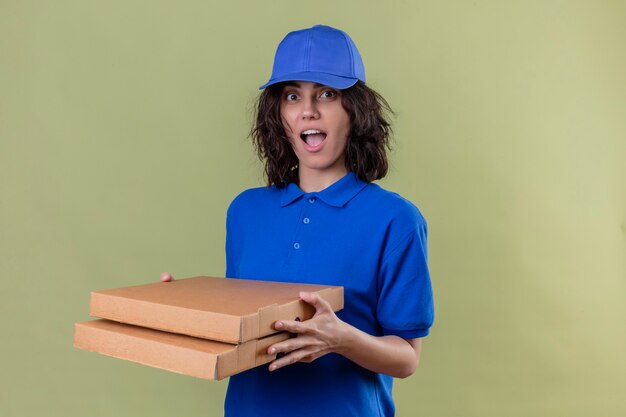 Liefermädchen in der blauen Uniform und in der Kappe, die Pizzaschachteln halten, die freudig positiv und glücklich lächelnd fröhlich über isolierten Grünflächen stehend schauen