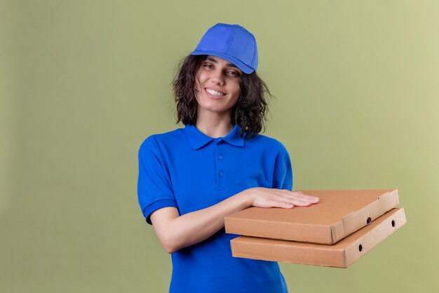 Liefermädchen in der blauen Uniform und in der Kappe, die Pizzaschachteln positiv und glücklich lächelnd freundlich stehen auf isoliertem Grün hält