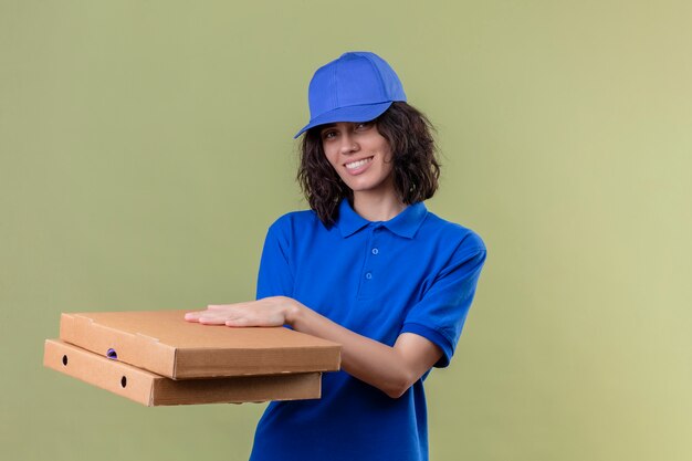 Liefermädchen in der blauen Uniform und in der Kappe, die Pizzaschachteln positiv und glücklich lächelnd freundlich stehen auf isoliertem Grün hält