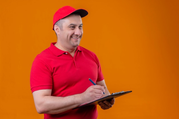 Liefermann, der rote Uniform und Kappe hält, die Zwischenablage hält, die beiseite positiv und glücklich schaut, etwas schreibend über orange Wand