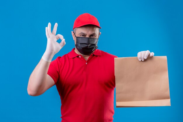 Liefermann, der rote Uniform und Kappe in der Gesichtsschutzmaske trägt, die Papierpaket positiv und glücklich hält, OK Zeichen über blaue Wand zu tun