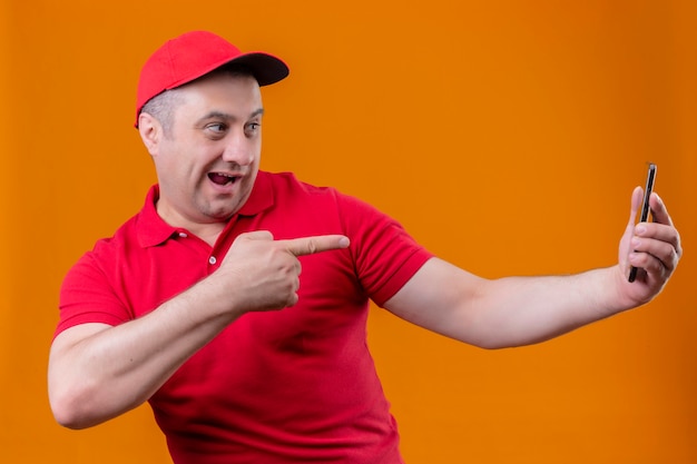 Liefermann, der rote Uniform und Kappe trägt, die freudig hält, Smartphone hält und mit Zeigefinger darauf zeigt, der über orange Wand lächelt