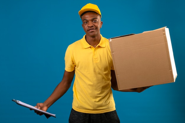 Lieferung Afroamerikaner Mann in gelbem Poloshirt und Kappe, die Klemmbrett und Pappkarton lächelnd freundlich stehend auf isoliertem Orange hält