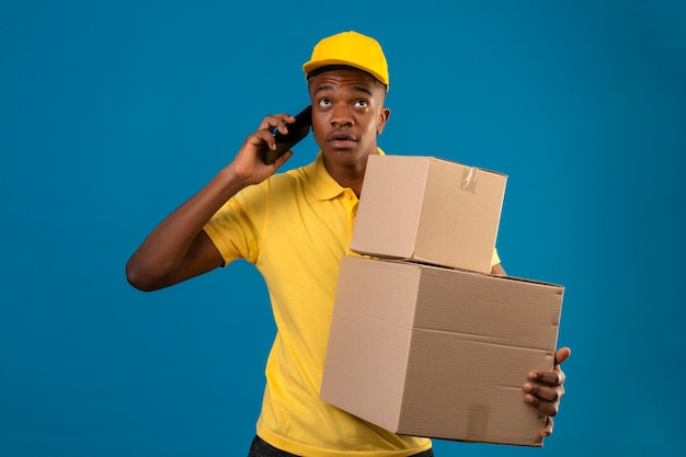 Lieferung afroamerikaner mann in gelbem poloshirt und kappe stehend mit pappkartons, die auf mobiltelefon sprechen, das nachdenklichen blick auf lokalisiertem blau sucht