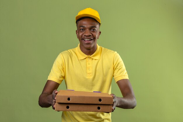 Lieferung Afroamerikaner Mann in gelbem Poloshirt und Kappe stehend mit Pizzaschachteln lächelnd freundlich mit glücklichem Gesicht, das auf Grün steht