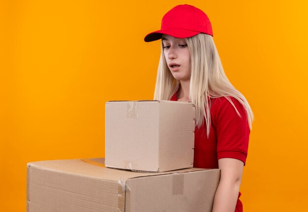 Lieferung junge Frau, die rotes T-Shirt und Kappe hält, die Kisten auf isolierter orange Wand hält