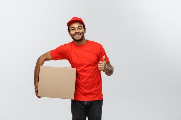 Lieferung Konzept - Porträt von Happy African American Lieferung Mann mit Box-Pakete und zeigt thumps up. Isoliert auf grau studio Hintergrund. Text kopieren