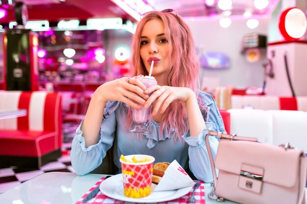 Lifestyle-Innenbild der stilvollen jungen hübschen Frau mit welligen ungewöhnlichen rosa Haaren und natürlichem Make-up, niedliches blaues Kleid und Jeansjacke tragend, genießen Sie ihr leckeres amerikanisches Abendessen.