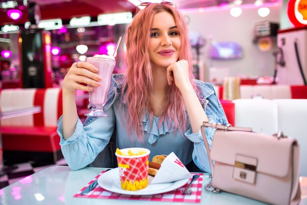 Lifestyle-Innenbild der stilvollen jungen hübschen Frau mit welligen ungewöhnlichen rosa Haaren und natürlichem Make-up, niedliches blaues Kleid und Jeansjacke tragend, genießen Sie ihr leckeres amerikanisches Abendessen.