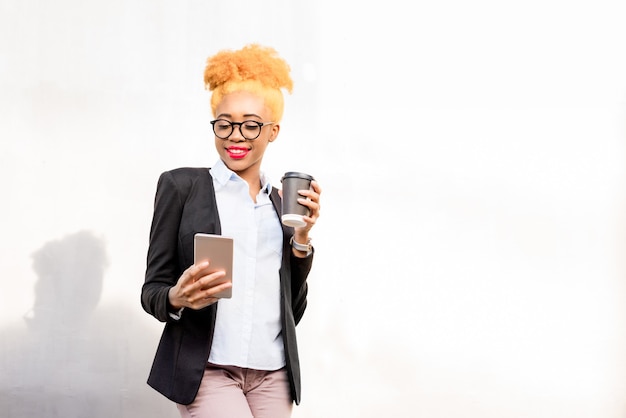 Lifestyle-porträt einer afrikanischen geschäftsfrau in brille und freizeitanzug, die mit telefon und kaffeetasse auf dem grauen wandhintergrund steht