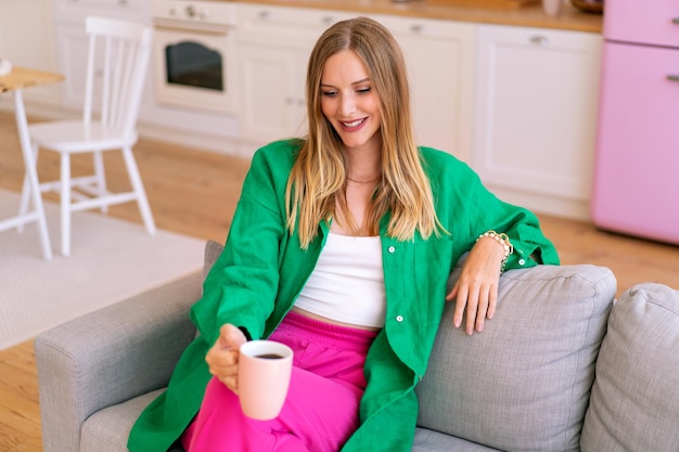 Lifestyle-porträt einer stilvollen blonden frau entspannt auf ihrem sofa, trägt grünes leinenhemd und fucsiafarbene hosen und trinkt morgenkaffee.