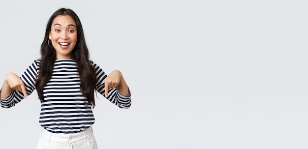 Lifestyle Schönheit und Mode Menschen Emotionen Konzept Aufgeregt zart charmantes asiatisches Mädchen mit süßem Lächeln, das mit dem Finger nach unten zeigt und Kunden zu einem besonderen Ereignis auf weißem Hintergrund einlädt