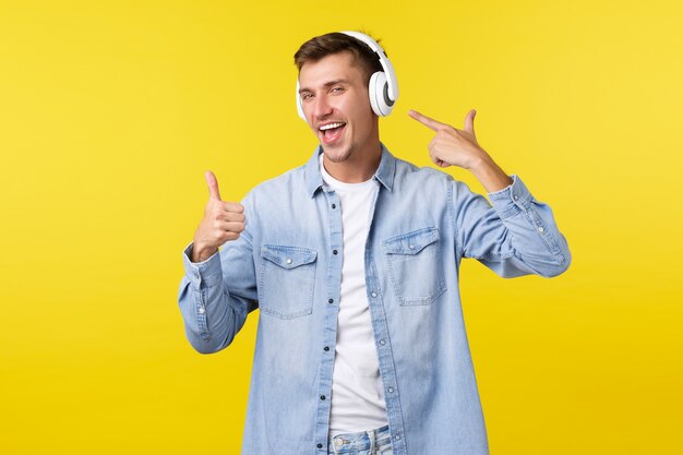 Lifestyle, Sommerferien, Technologiekonzept. Fröhlicher gutaussehender Mann, Student in drahtlosen Kopfhörern, der auf Kopfhörer zeigt und Daumen hoch zeigt, als zufrieden mit guter Musik, tollen Beats.