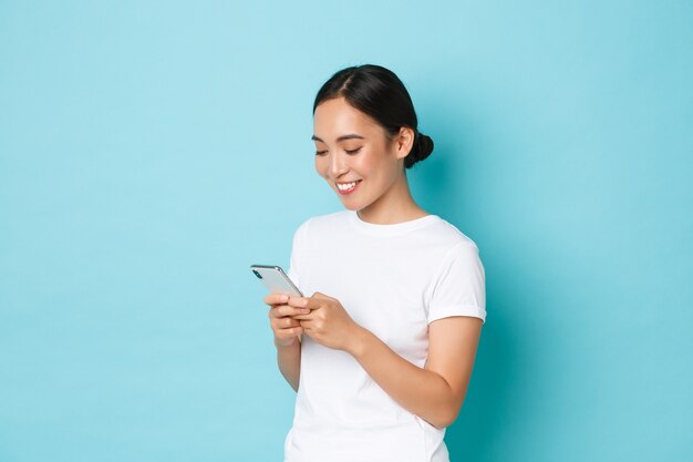 Lifestyle-, Technologie- und E-Commerce-Konzept. Seitenansicht des attraktiven asiatischen Mädchens, das Handy, SMS, Nachrichten oder Chat mit Freunden online verwendet, Blick auf Smartphone-Bildschirm erfreut.