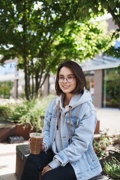 Lifestyle-Wochenenden und People-Konzept Porträt einer jungen kaukasischen Frau in einer Jeansjackenbrille, die eine glückliche Kamera sieht, während sie auf einer Parkbank mit Kaffeetrinken-Eislatte sitzt und den Frühling genießt