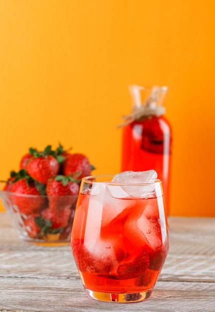 Limonade mit Erdbeeren in Krug und Glas auf Holz- und Orangenwand, Seitenansicht.