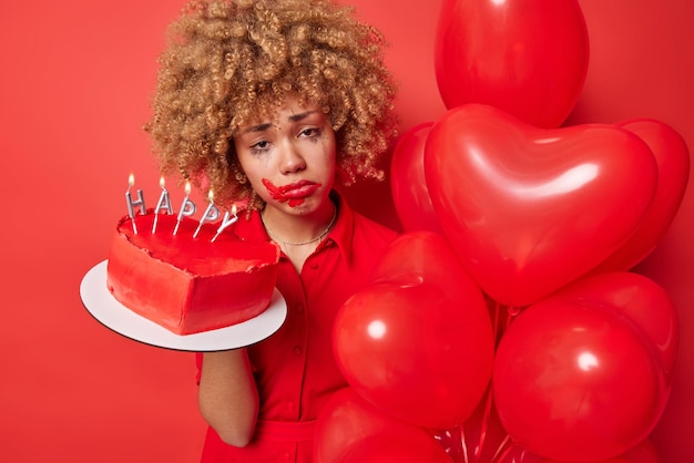 Lockige behaarte Frau hat verwöhntes Make-up, sieht frustriert aus, hat einen unglücklichen Ausdruck, fühlt sich während des Urlaubs einsam an und hält süßen Kuchen und herzförmige Luftballons, die über rotem Hintergrund isoliert sind Festkonzept