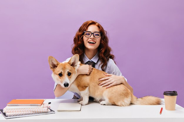 Lockige Dame in Hemd und Brille lächelt und spielt mit Corgi