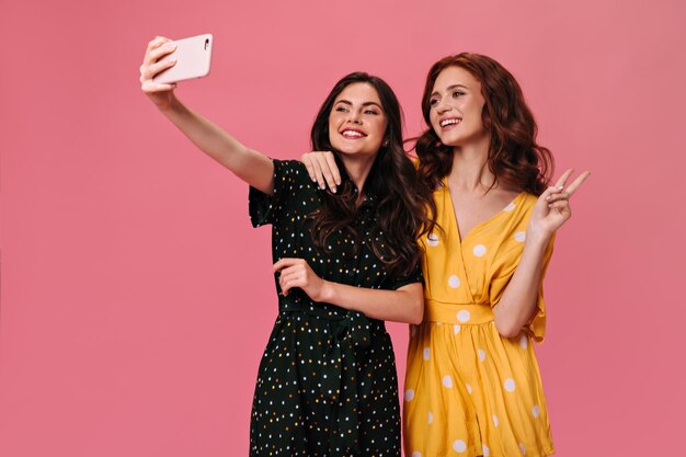 Lockige Damen in stylischen Sommerkleidern machen Selfie auf rosa Hintergrund Schöne Freundinnen in hellen Overalls posieren für Fotos