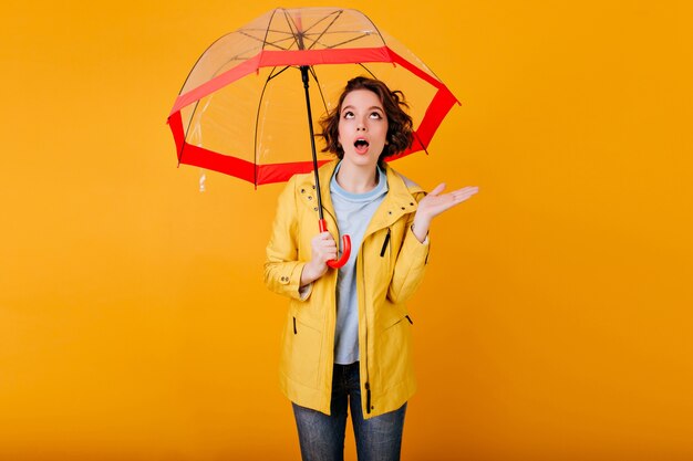 Lockige Frau im gelben Mantel, der Erstaunen ausdrückt, das unter Sonnenschirm steht. Porträt des emotionalen Mädchens mit Regenschirm, mit offenem Mund nach oben schauend.