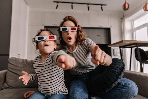 Lockige Frau und ihre Tochter sitzen mit 3D-Brille auf dem Sofa und schauen sich überrascht einen Film an