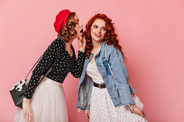 Lockige Mädchen im lässigen Outfit sprechen auf rosa Hintergrund. Studioaufnahme von raffinierten Freunden, die Gerüchte teilen.