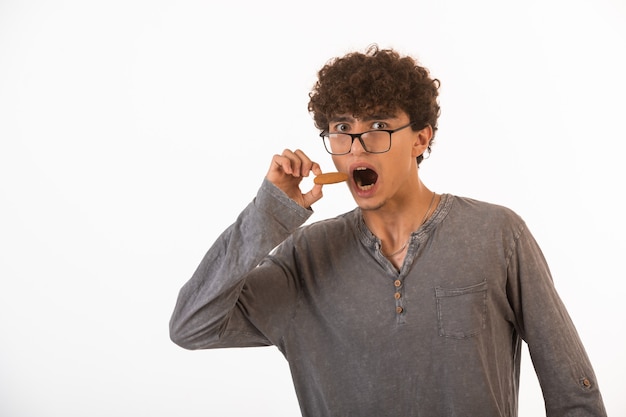 Lockiges haar junge in optischen gläsern weit öffnet seine augen und mund, um keks zu essen.